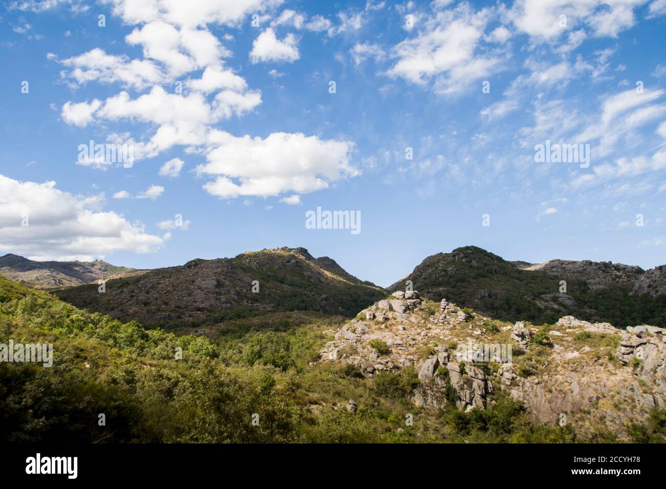 Mountain stone peaks stand uphill under a bright blue sky with white clouds hanging above Stock Photo
