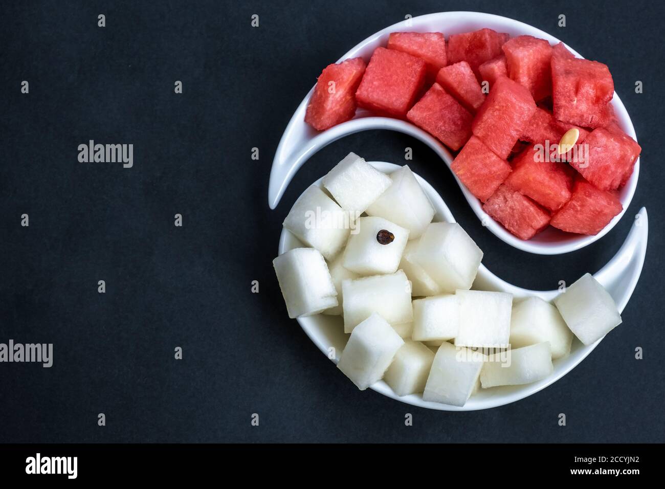 Ripe watermelon and melon, cut into cubes, are laid out on a yin-yang dish on a black background. Juicy, ripe summer fruits. Bright light. Copy space. Stock Photo