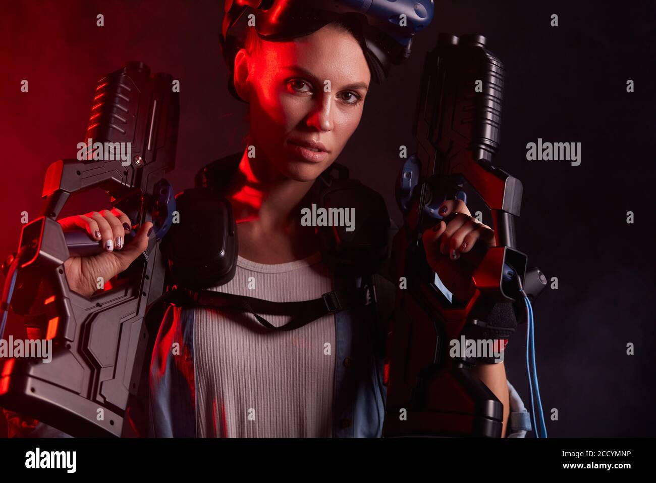 Portrait of beautiful woman holding weapons for virtual reality game, isolated over black background with red backlit. Look at camera Stock Photo