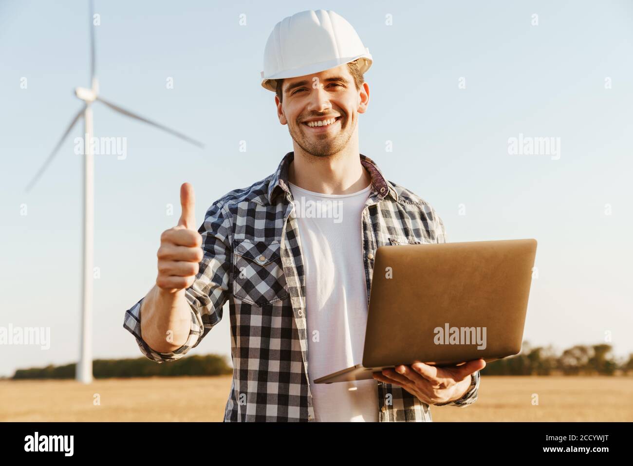 An electrical engineer standing on a field with windmills, using laptop computer, thumbs up Stock Photo