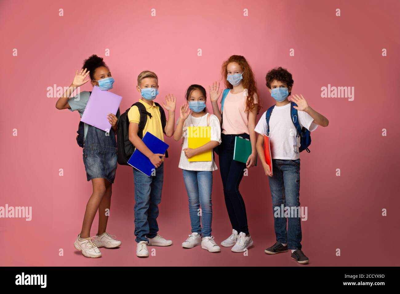 Education during virus pandemic. Full length portrait of schoolkids in medical masks waving on pink background Stock Photo