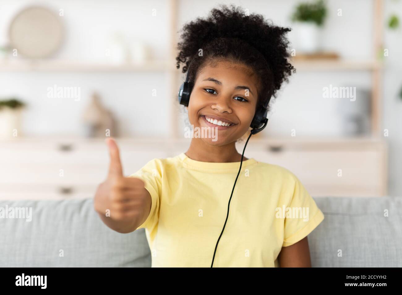 Black Girl With Headphones Gesturing Thumbs-Up Sitting On Couch Indoor Stock Photo