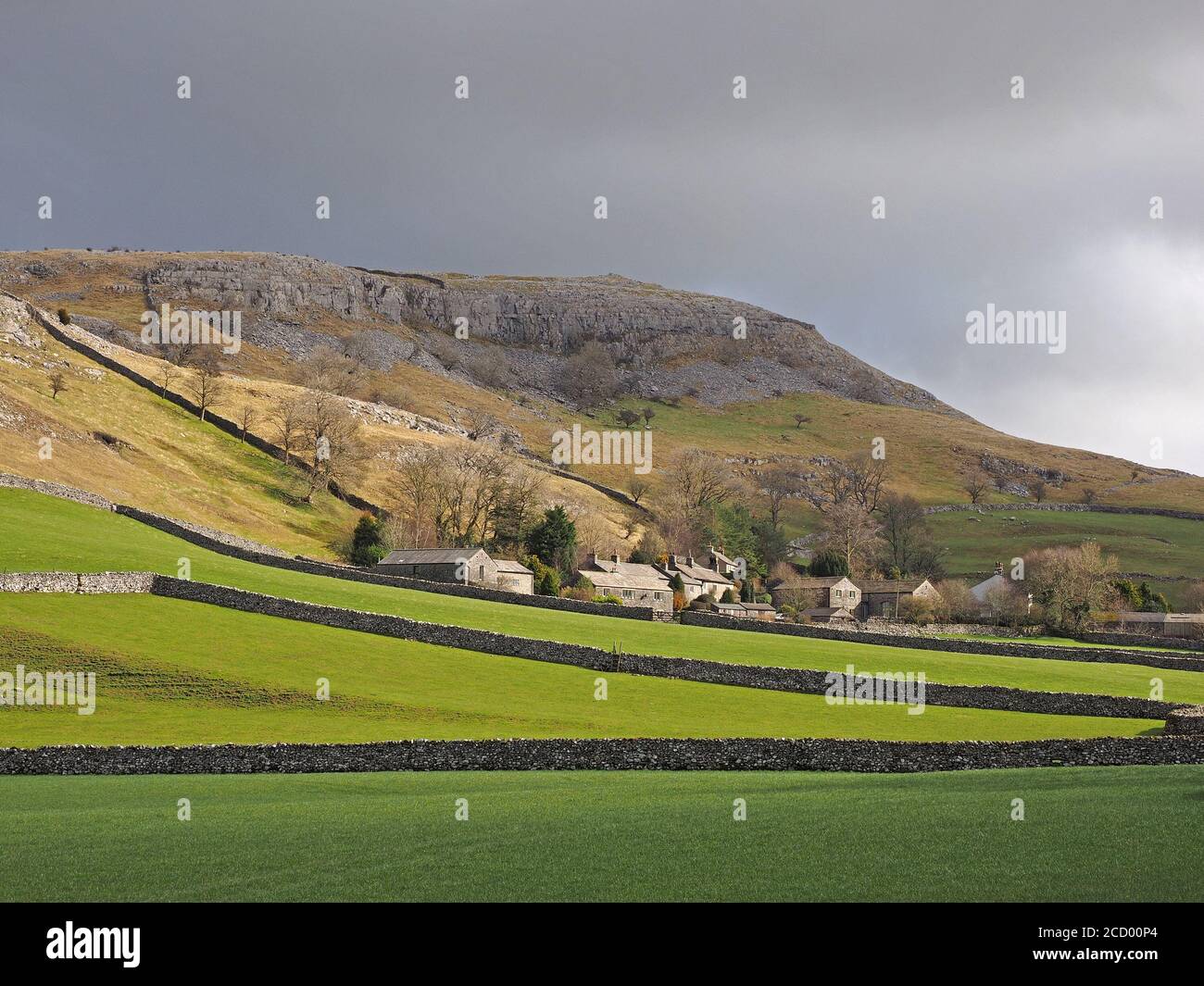 view of stone buildings of Feizor village amid green fields, dry-stone walls below craggy limestone scar nr Austwick, North Yorkshire, England, UK Stock Photo