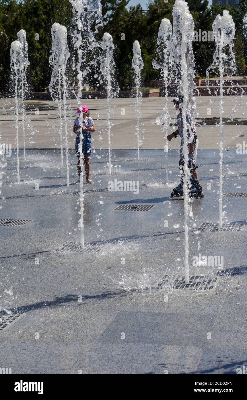 Nikolaev, Ukraine - August 17, 2020: Children rollerblading among the jets of the fountain. Water jets of the fountain in the city square. Jets of coo Stock Photo