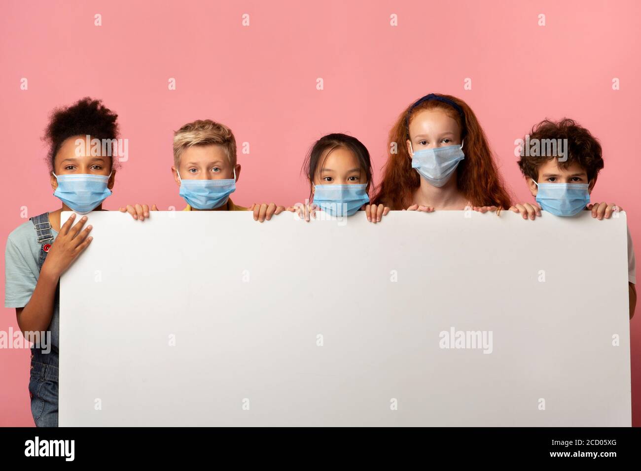 Group of schoolkids in protective masks holding empty banner with space for design over pink background Stock Photo