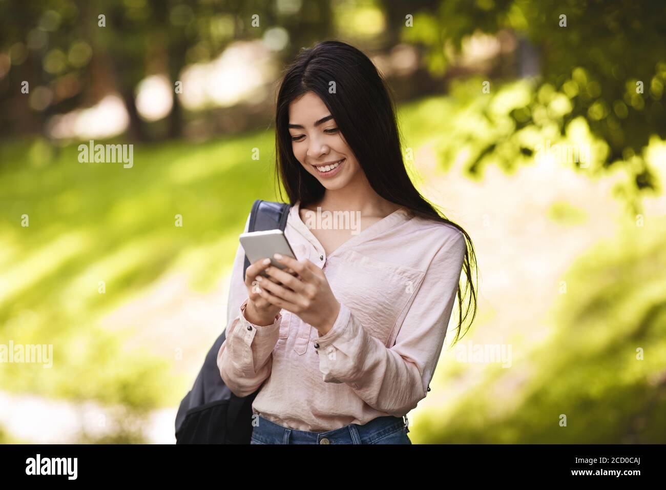 Cheerful asian teen student girl with backpack using smartphone outdoors Stock Photo