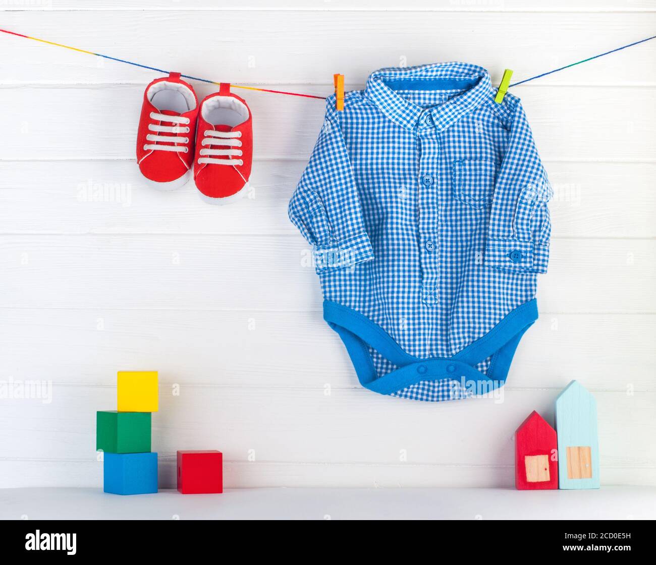 Red baby booties and blue bodykit on clothespins and wooden toys over white wooden background with blank space for text. Stock Photo