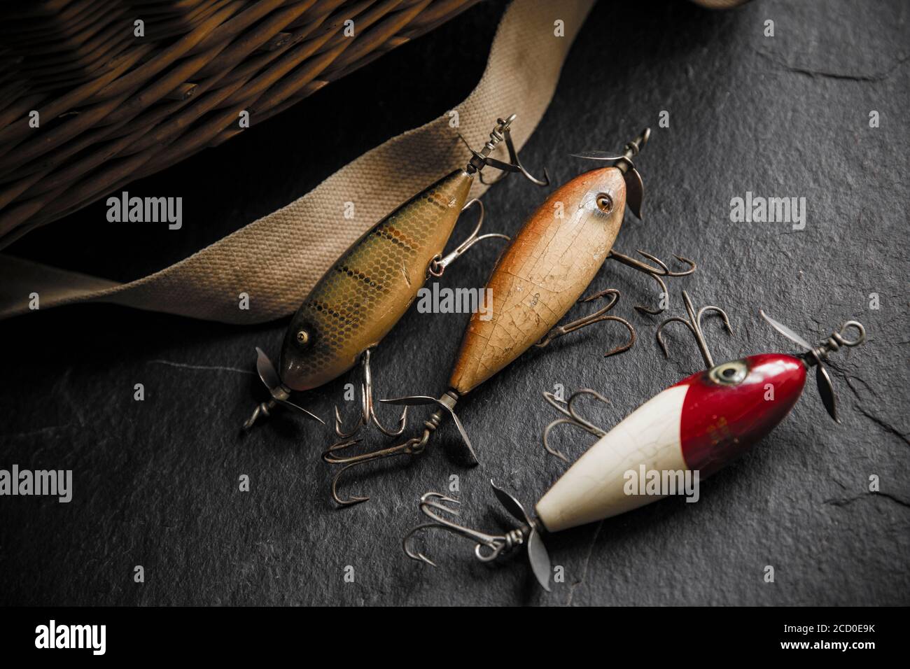 Examples of old South Bend fishing lures, or plugs, designed to catch predatory fish displayed on a dark slate background next to an old whicker tackl Stock Photo
