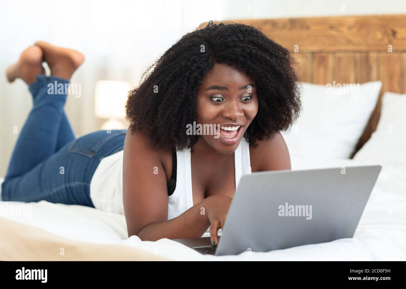 Surprised black girl lying on bed, using laptop Stock Photo - Alamy