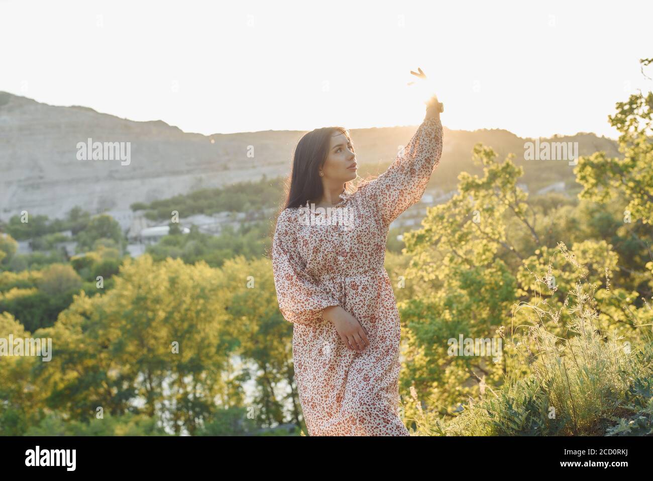 The young woman is catching the sun rays. The young woman is on the hill. The brunette is in the sunlight. Stock Photo