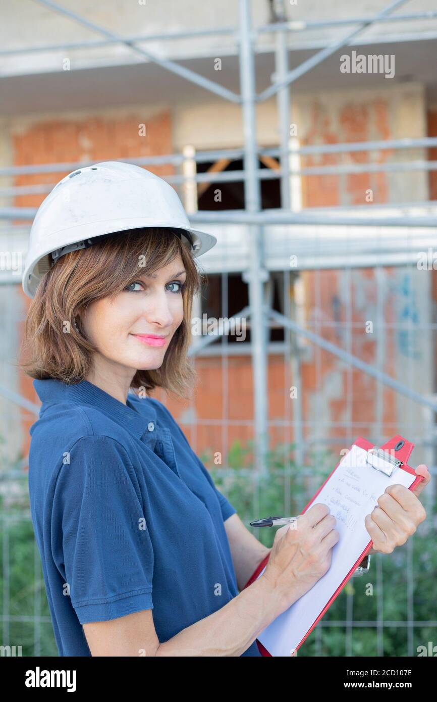 Woman, female engineer, caucasian, age 40, wearing a safety white cap, looking at camera, working on a costruction site in a typical men's role. Gende Stock Photo