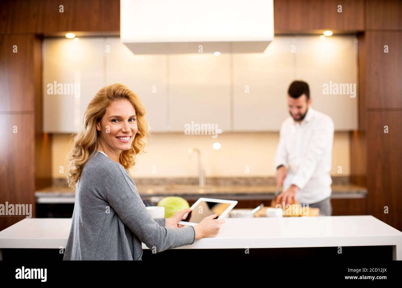 Young woman using tablet at the kitchen table while man preparing food Stock Photo