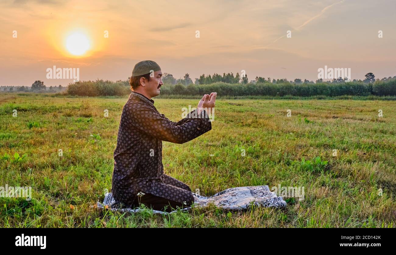 A Muslim senior man wearing a skullcap and traditional clothes prays at sunset Stock Photo