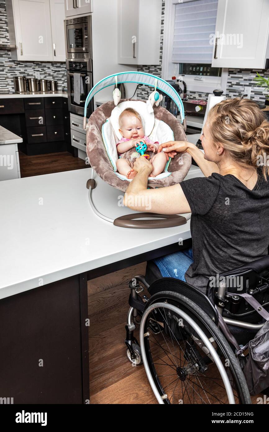 A paraplegic mother in a wheelchair playing with her baby in a baby seat, while in her kitchen; Edmonton, Alberta, Canada Stock Photo