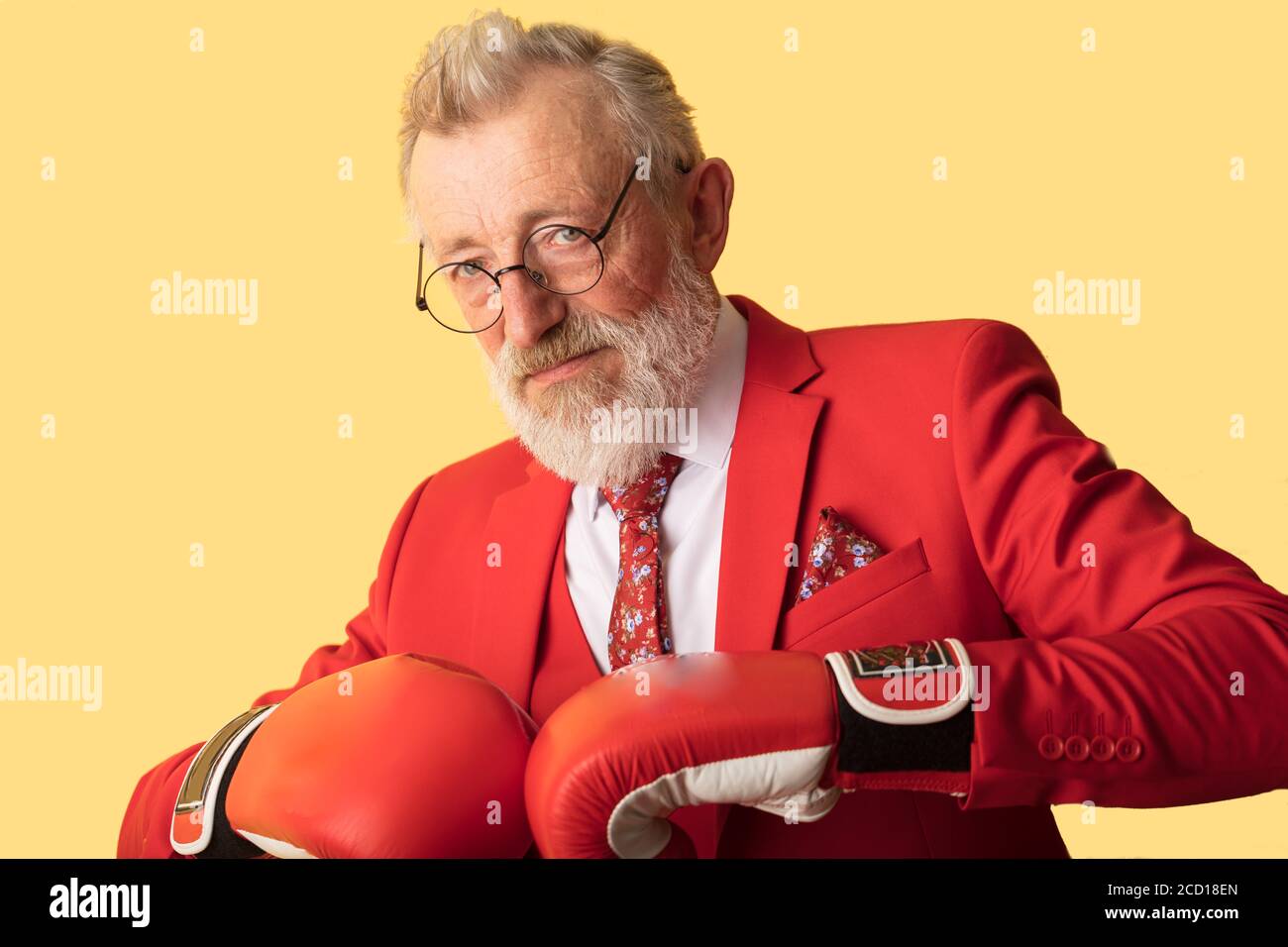 Senior business man wearing red formal suit and red boxing gloves looking at camera while standing in defence pose on yellow background. Economic comp Stock Photo