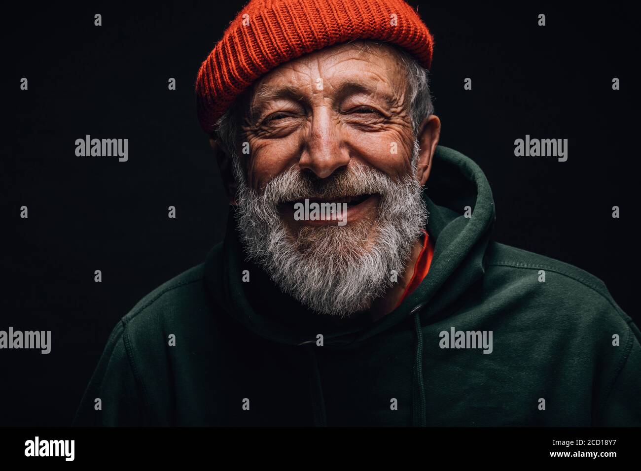 Close up portrait of happy 70-year-old optimist man with smiling wrinkled face, dressed in hipster orange hat and green hoodie, isolated over black ba Stock Photo