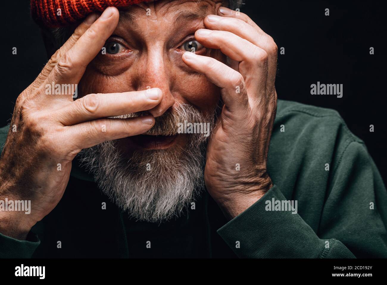 An elderly bearded man looking extremely surprised or frightened, protecting his face with hands, close up face portrait over black studio background. Stock Photo