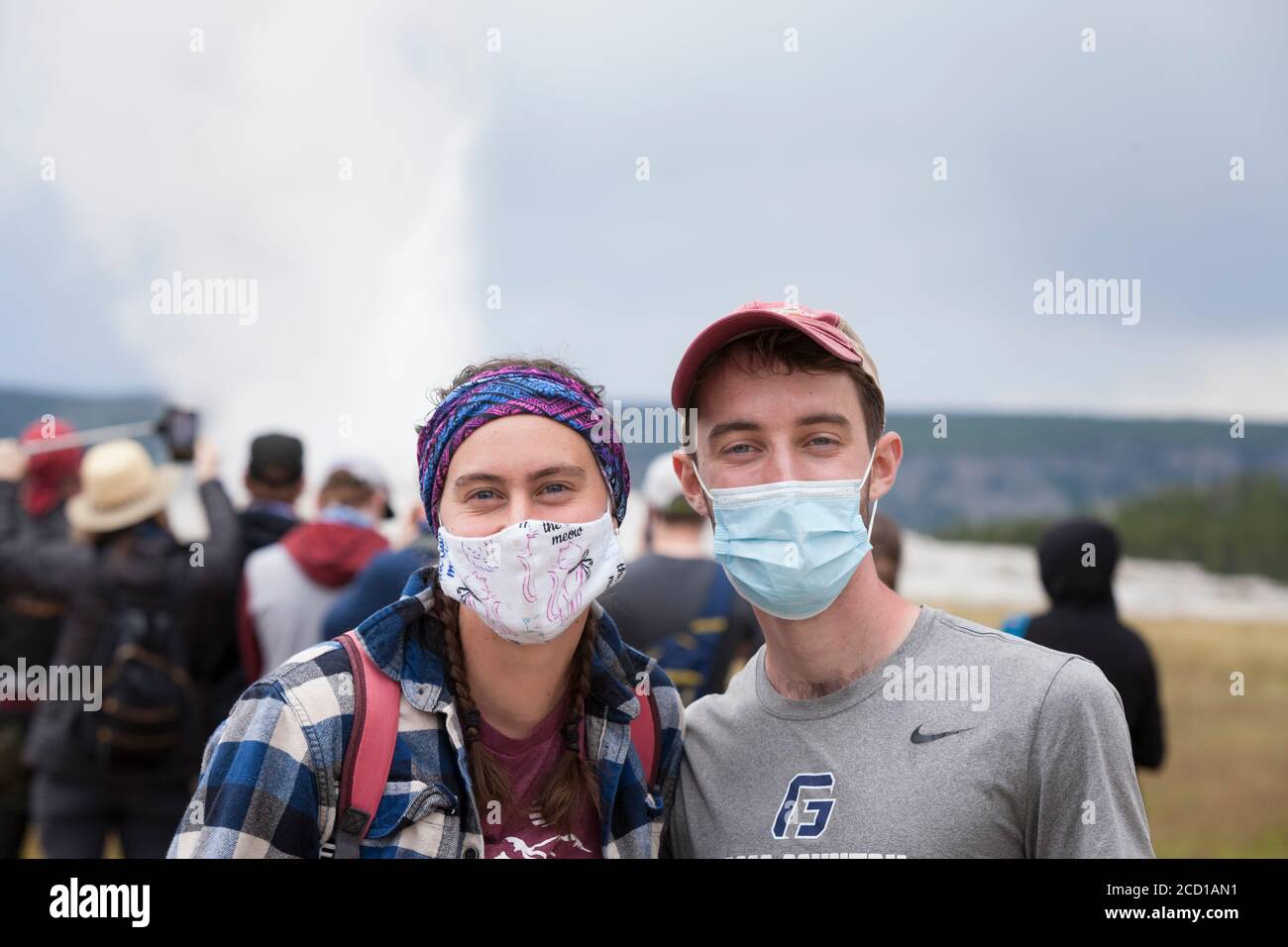 A young couple from Oregon wearing face masks pose for a photo as visitors clamor for photos of Old Faithful erupting at Yellowstone National Park, Wy Stock Photo