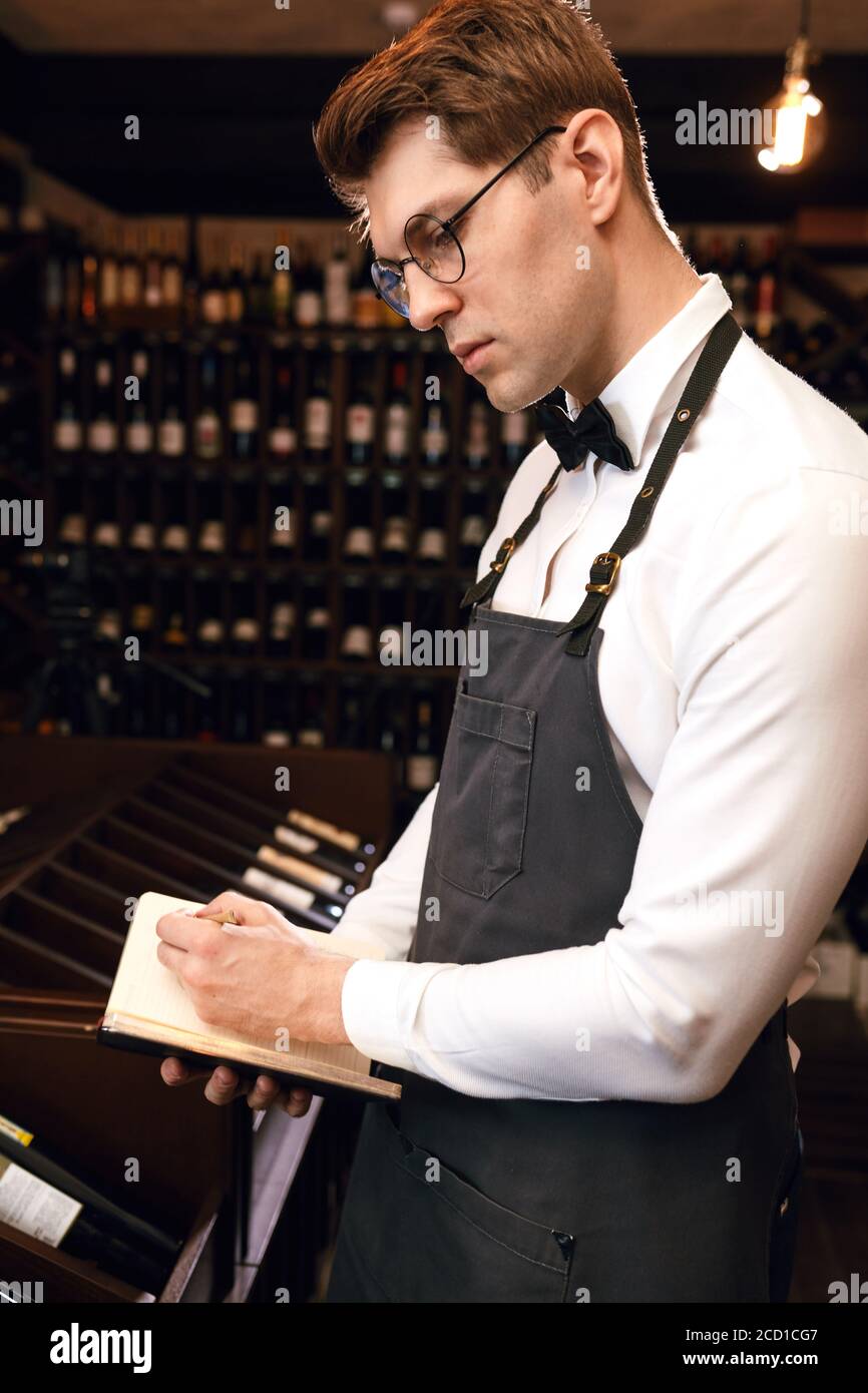 Handsome male wine steward standing with notepad against long rows of shelves with wine bottles at wine store Stock Photo