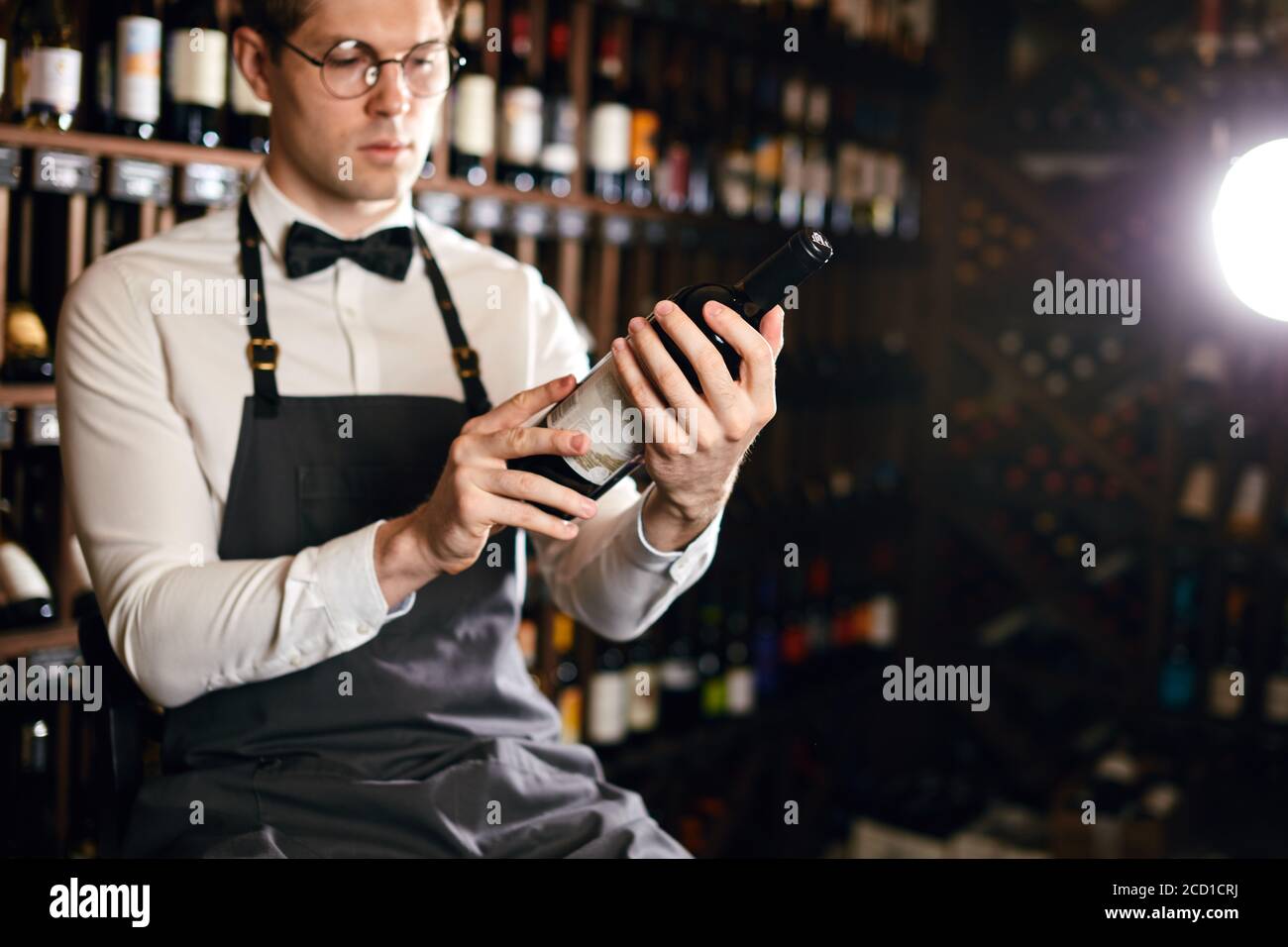 Young caucasian cavist dressed in white shirt and bowtie working in big vine shop presenting a bottle of red wine to customer Stock Photo