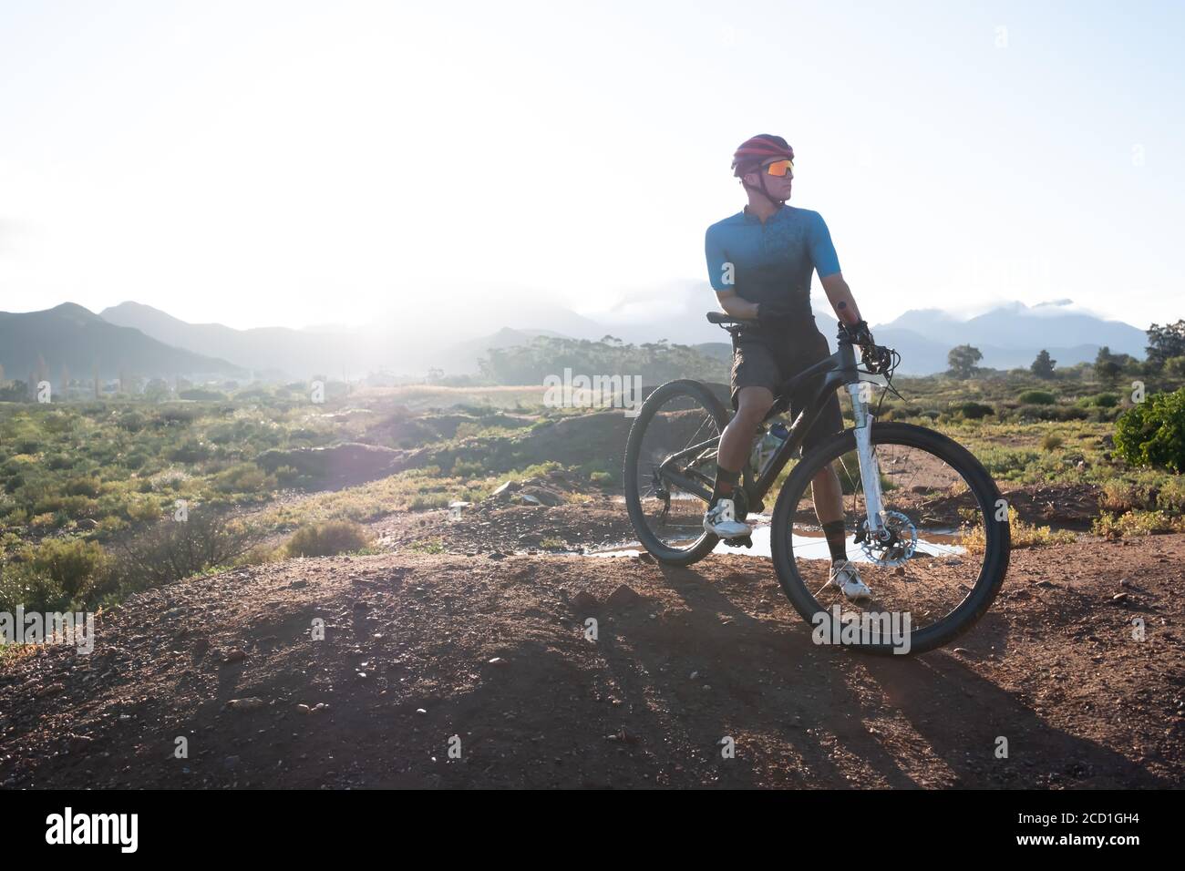 backlit image of a mountain biker and his bike on top of a mound with a mountain range in the distance Stock Photo