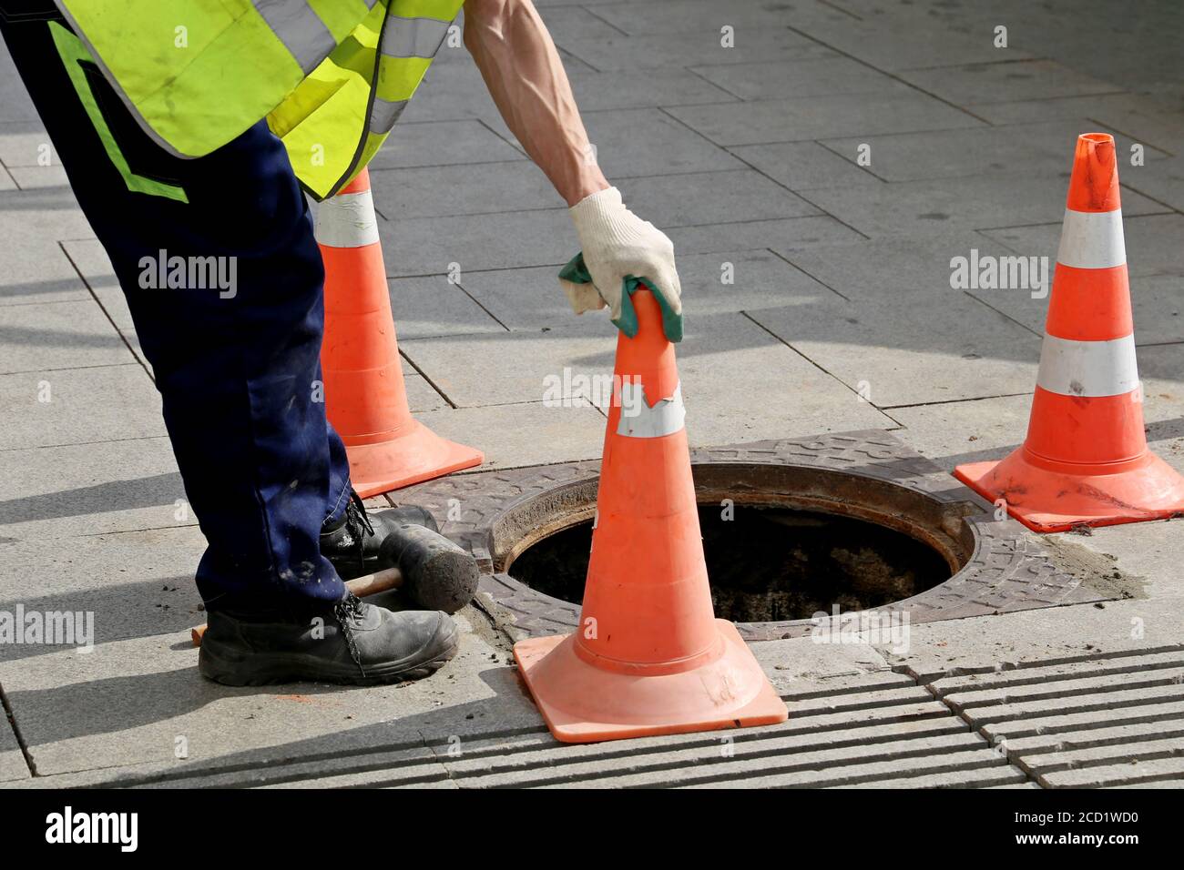 Worker over the open sewer hatch on a street near the traffic cones. Concept of repair of sewage, underground utilities, water supply system Stock Photo