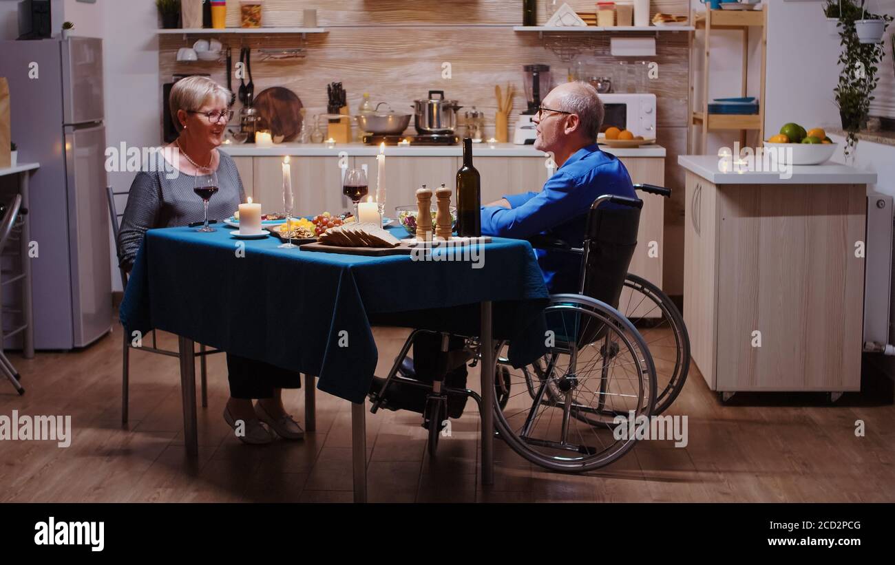 Senior couple coming home for romantic dinner. Old man in wheelchair dining with his cheerful wife sitting at the table in the kitchen. Imobilized paralyzed handicapped husband having romantic dinner Stock Photo