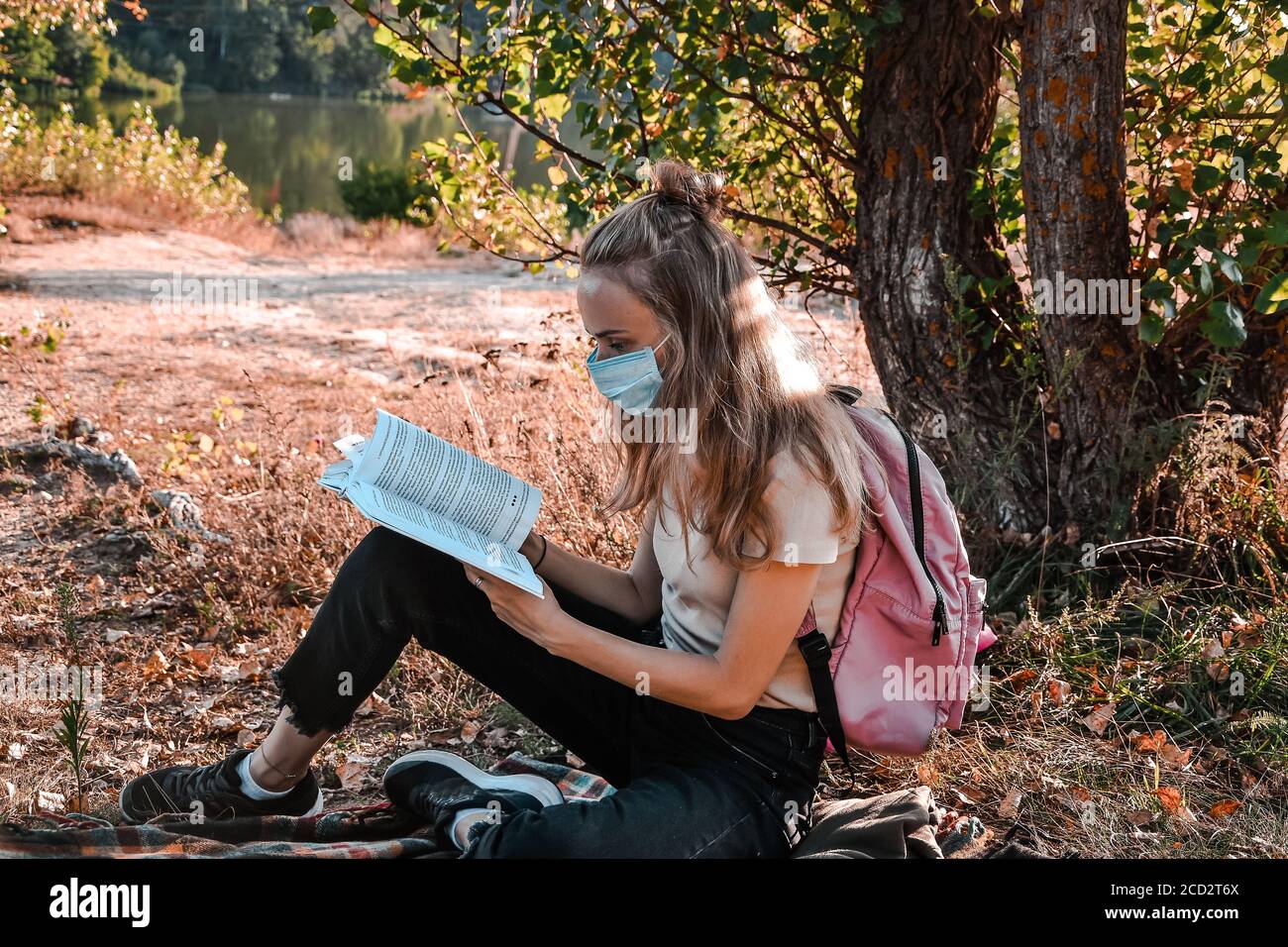 Young girl, student sitting outdoor with backpack and reading a book. Wearing protective face mask. Study from home. Cottagecore Stock Photo