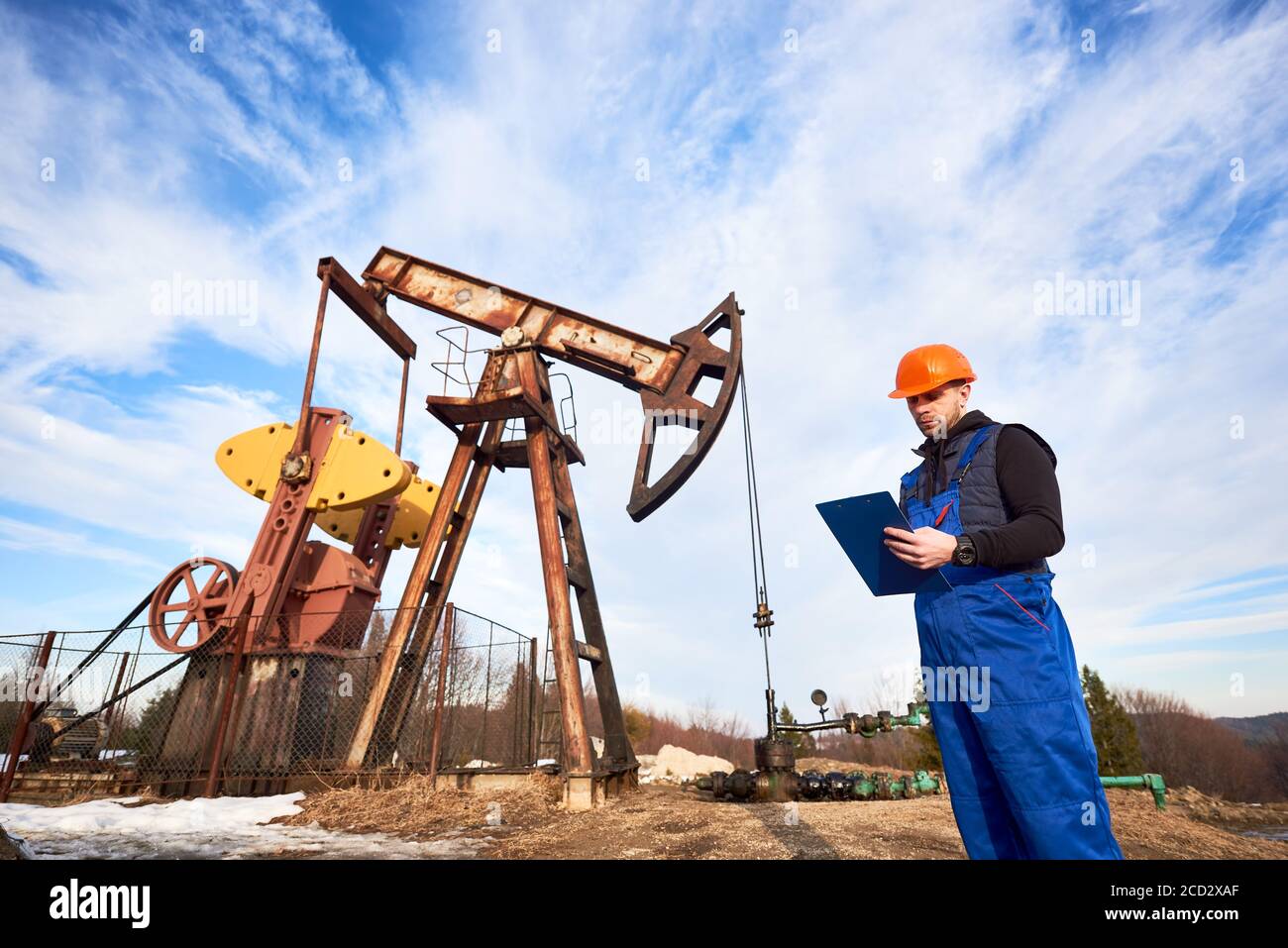 Petroleum engineer in work overalls and helmet holding clipboard, checking oil pumping unit, making notes. Oil worker standing near oil pump jack under beautiful sky. Concept of oil extraction Stock Photo