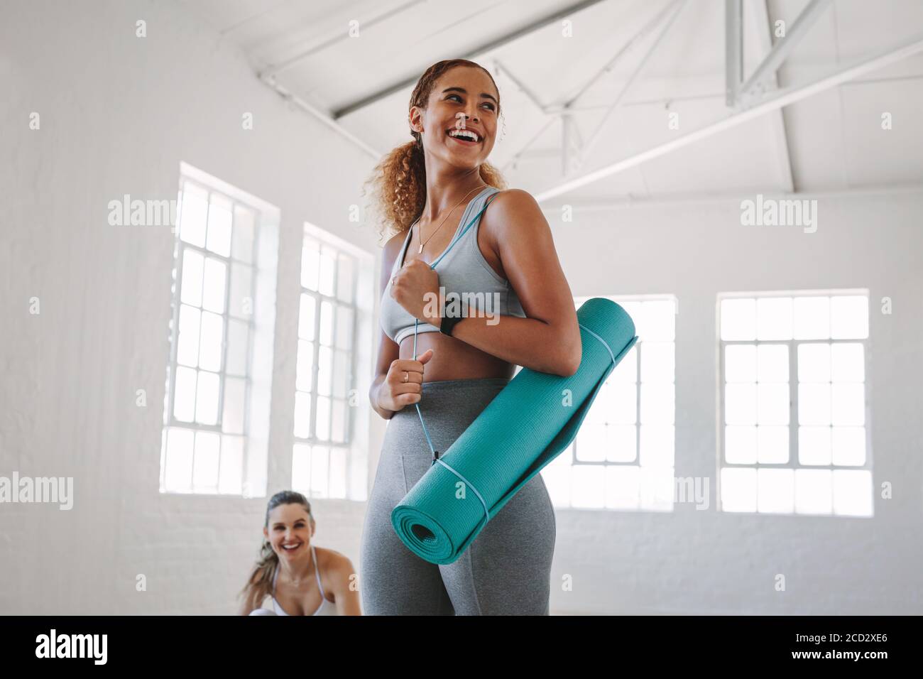 Smiling fitness woman standing in a fitness studio carrying a yoga mat. Portrait of a young woman at a fitness training centre. Stock Photo
