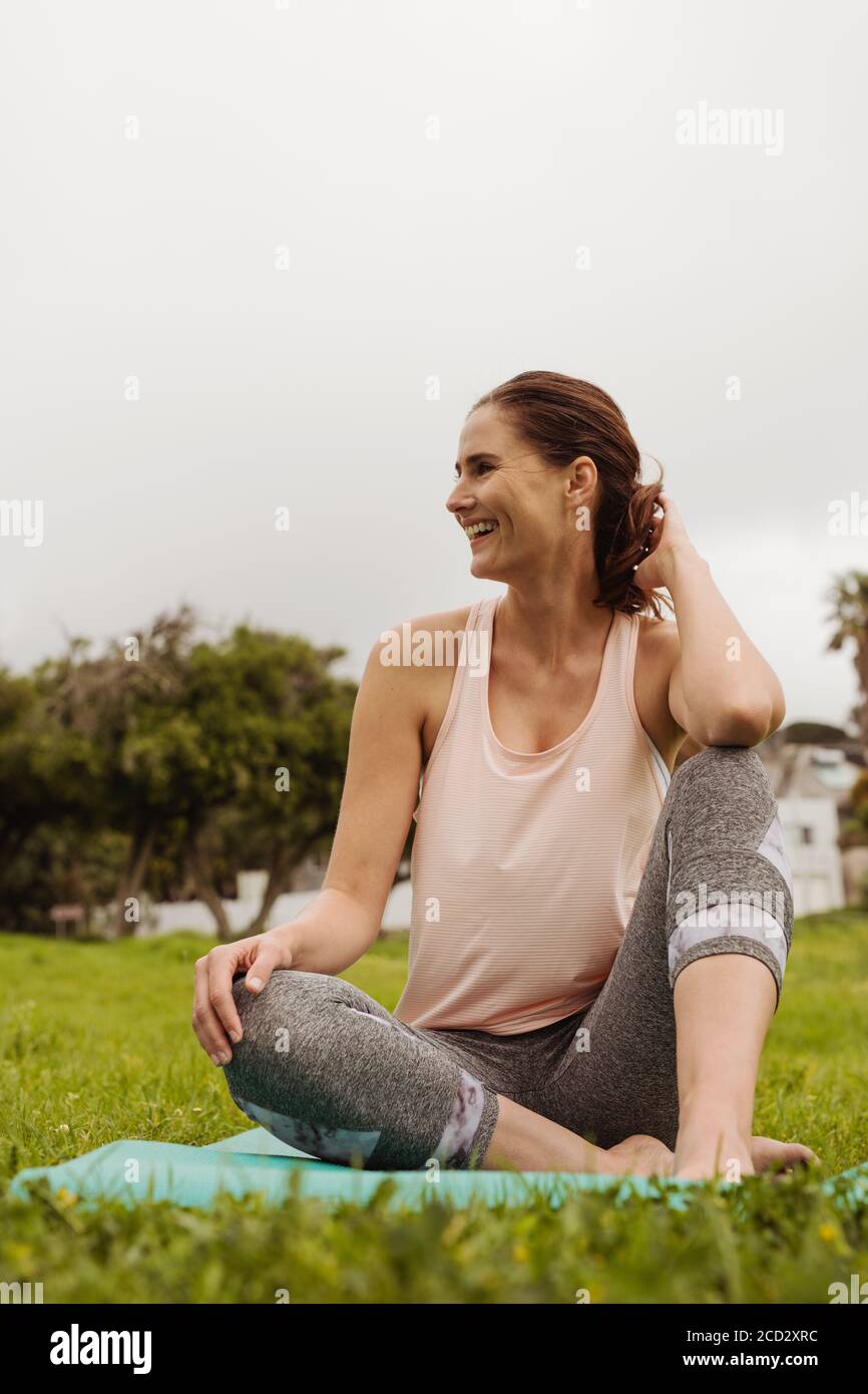 Portrait of a smiling woman sitting in park on a fitness mat. Cheerful female sitting in the meadows enjoying nature after her workout. Stock Photo