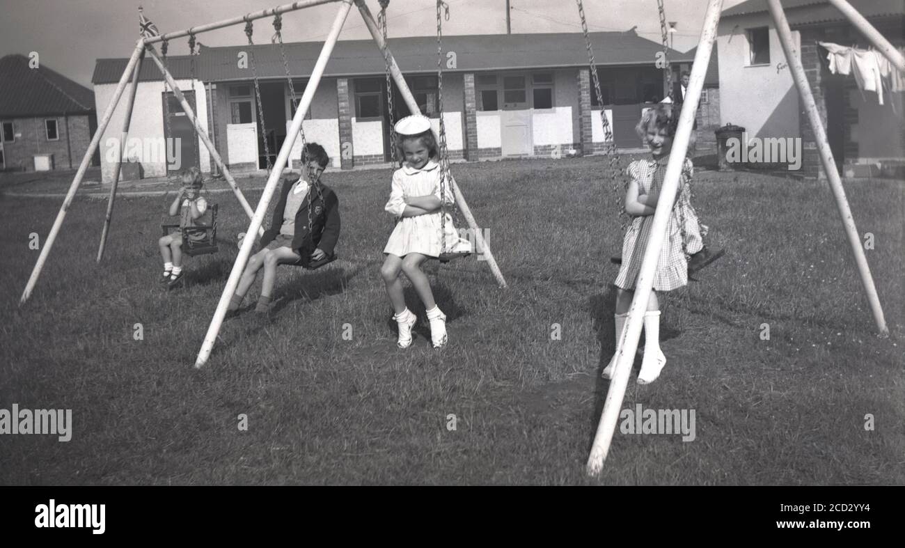 1954, historical, four young children, possibly all brothers and sisters, sitting outside on swings a grassy play area by a row of single-story chalets at a holiday camp on Hayling Island, Hampshire, England. Seaside holiday camps became very popular in post-war Britain during the 1950s and many were built to accomodate families who wanted an all-in type of holiday where everything was provided in one place. Stock Photo