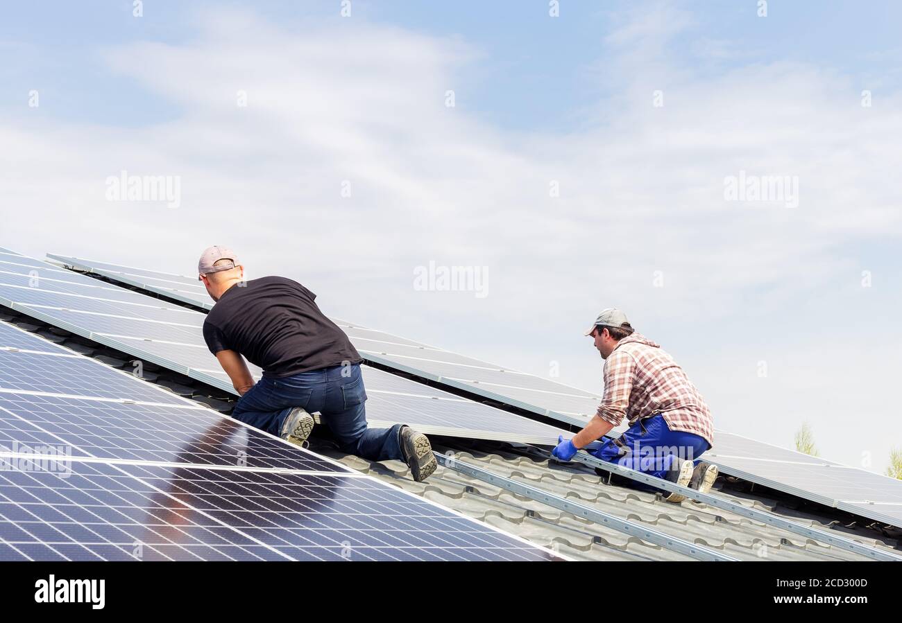 Process of building a solar station. Electrical engineers mans are working installing solar panels on solar station on house roof against blue sky Stock Photo