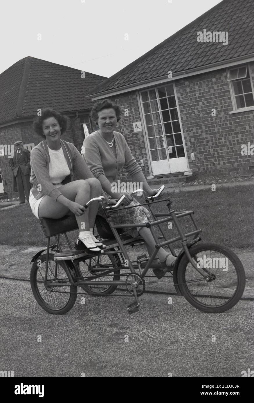 1950s, historical, two ladies sitting together on a tandem or two-seater social tricycle on a path outside a chalet at a holiday camp at Hayling Island, Hampshire, England, UK. These sturdy, three-wheel bicycles, where two friends or family members could sit together side-by-side whilist cycling, were a popular and fun form of transport to get around the large expanse of a holiday camp in this era. Stock Photo