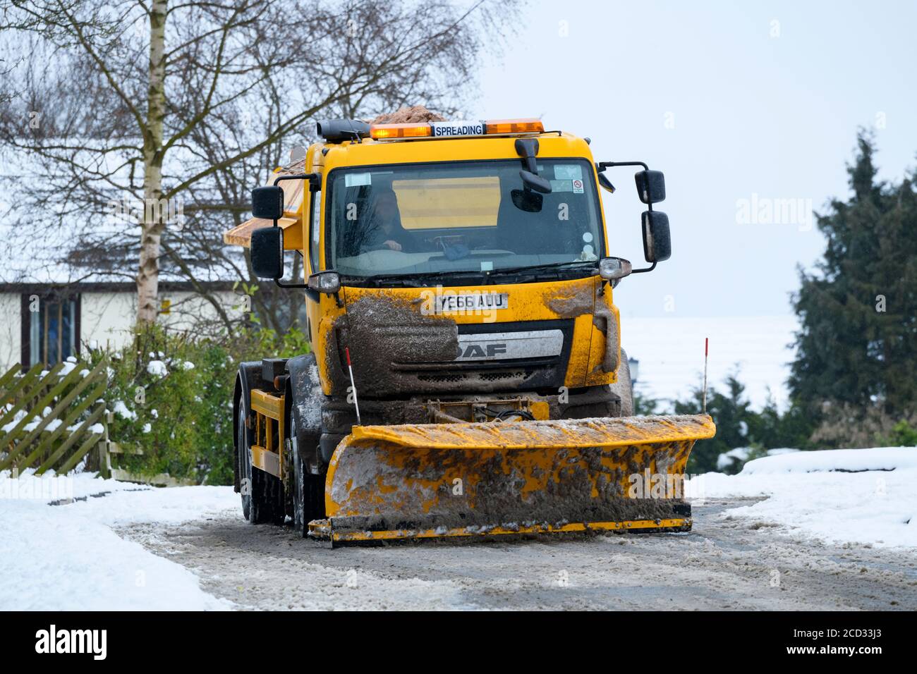 Cold snowy winter day (council gritter lorry, snow plough, man working & driving, spreading grit, clearing road) - Menston, West Yorkshire, England UK Stock Photo
