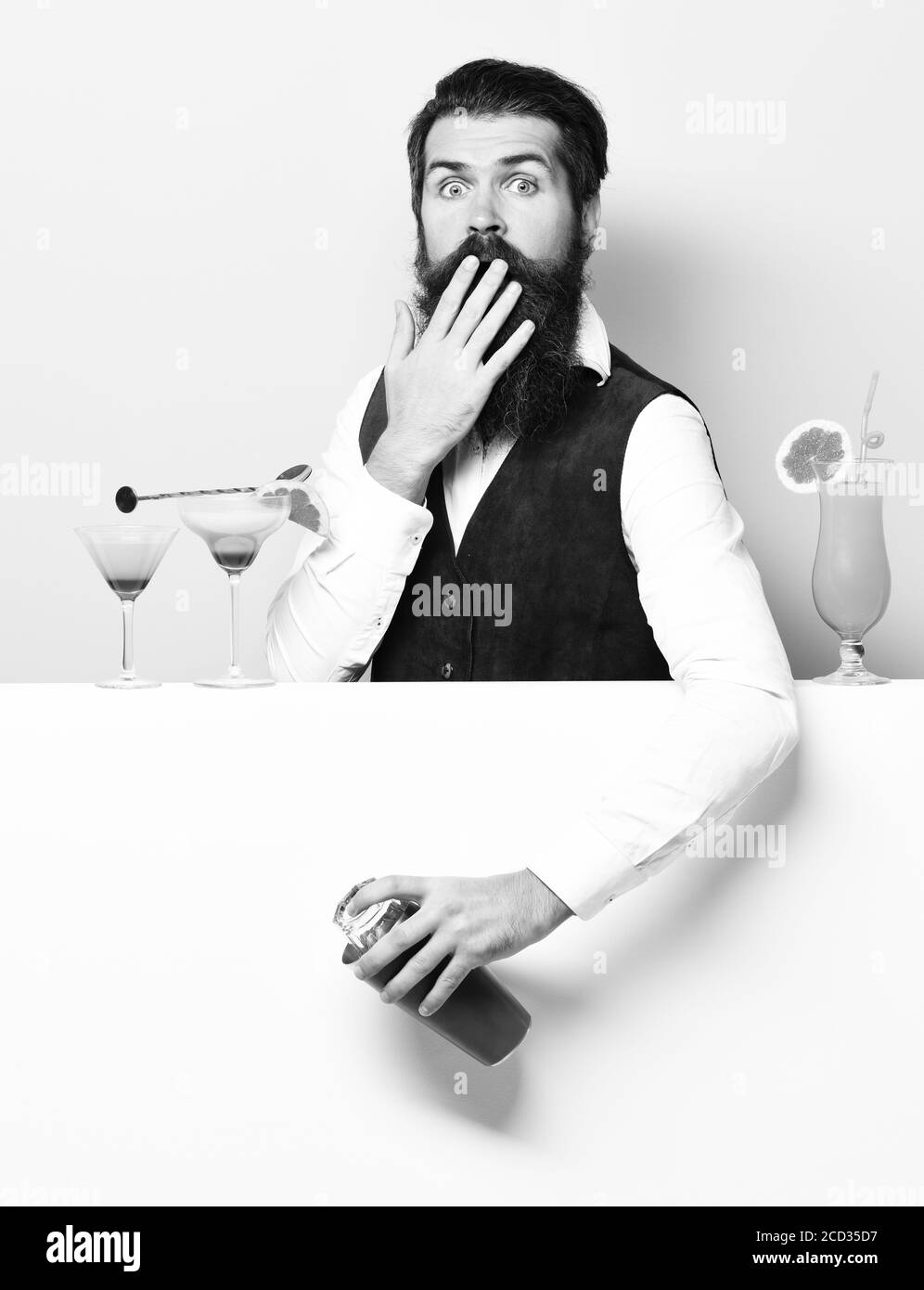 handsome bearded barman with long beard and mustache has stylish hair on surprised face holding shaker and made alcoholic cocktail in vintage suede leather waistcoat, on purple green studio background Stock Photo