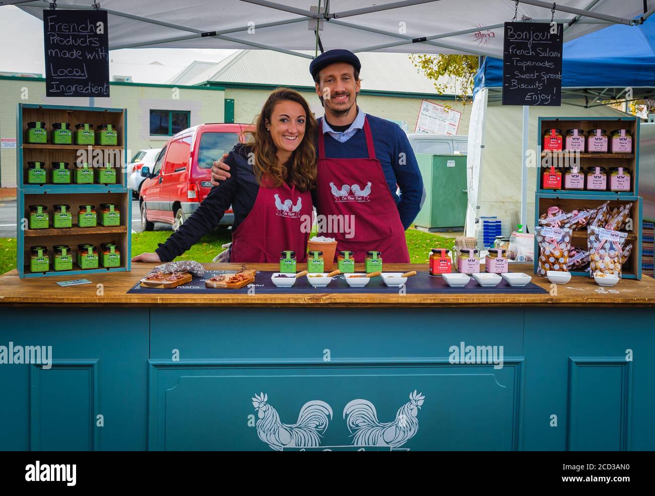 Young french couple in maroon aprons in front of their condiment stall at the Victor Harbor Farmers Markets in South Australia. Stock Photo