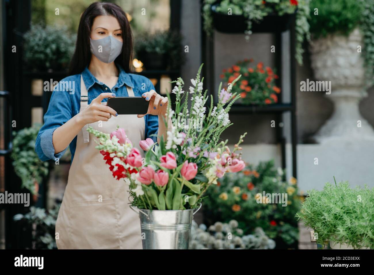 Florist at work. Woman in apron and protective mask makes photo on smartphone of bouquet Stock Photo