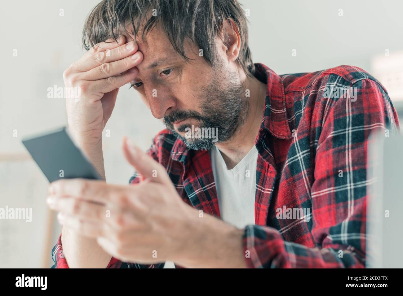 Troubled disappointed freelancer using smartphone for communication while sitting at the desk of his home office, selective focus Stock Photo