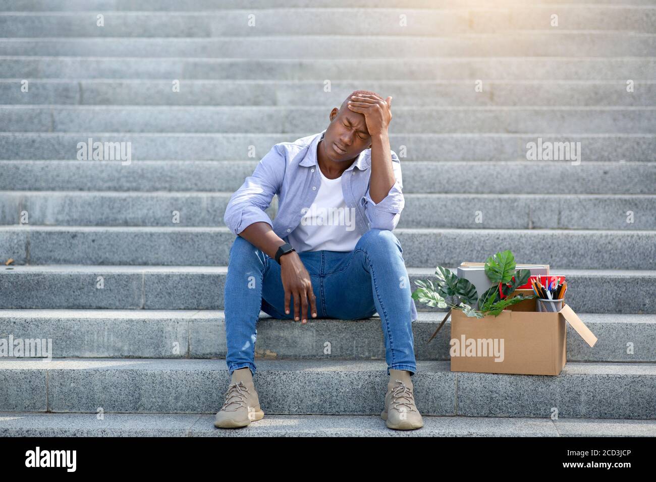 Depressed black guy with personal belongings sitting on stairs after losing his job, outdoors Stock Photo