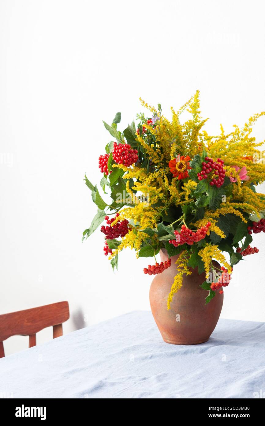 Beautiful bouquet of ragweeds, zinnia and branches of viburnum in a clay pot on a table against a white wall Stock Photo