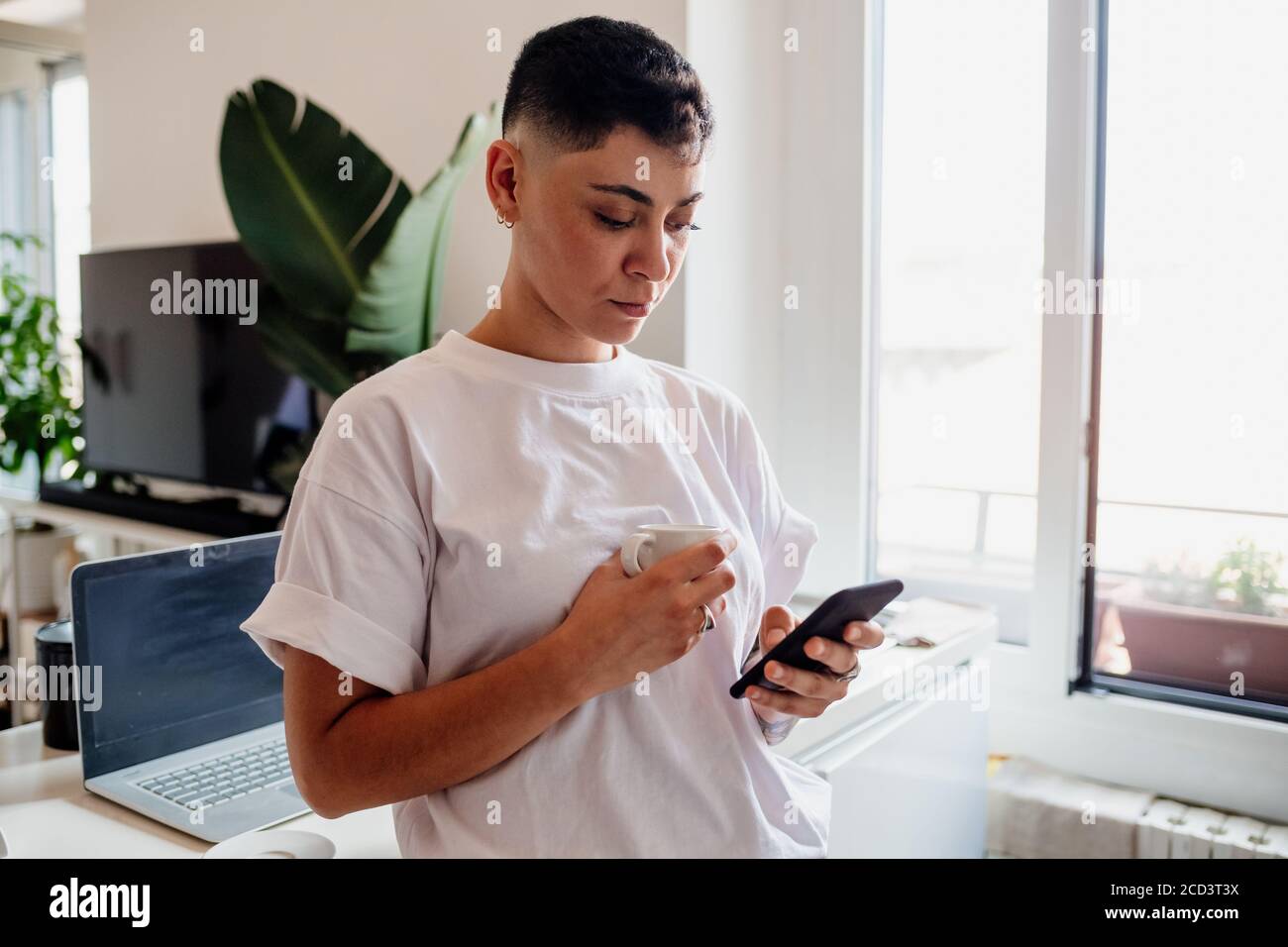Young woman with shaved head standing in a kitchen, checking her mobile phone. Stock Photo