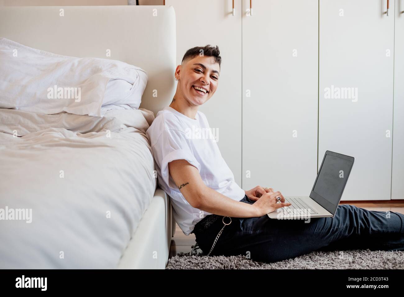 Young woman with shaved head sitting in bedroom, using laptop, smiling at camera. Stock Photo