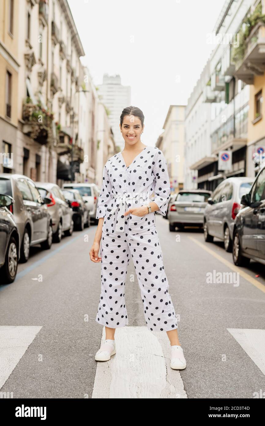 Portrait of smiling woman wearing white and black polka dot jumpsuit, standing on pedestrian crossing. Stock Photo