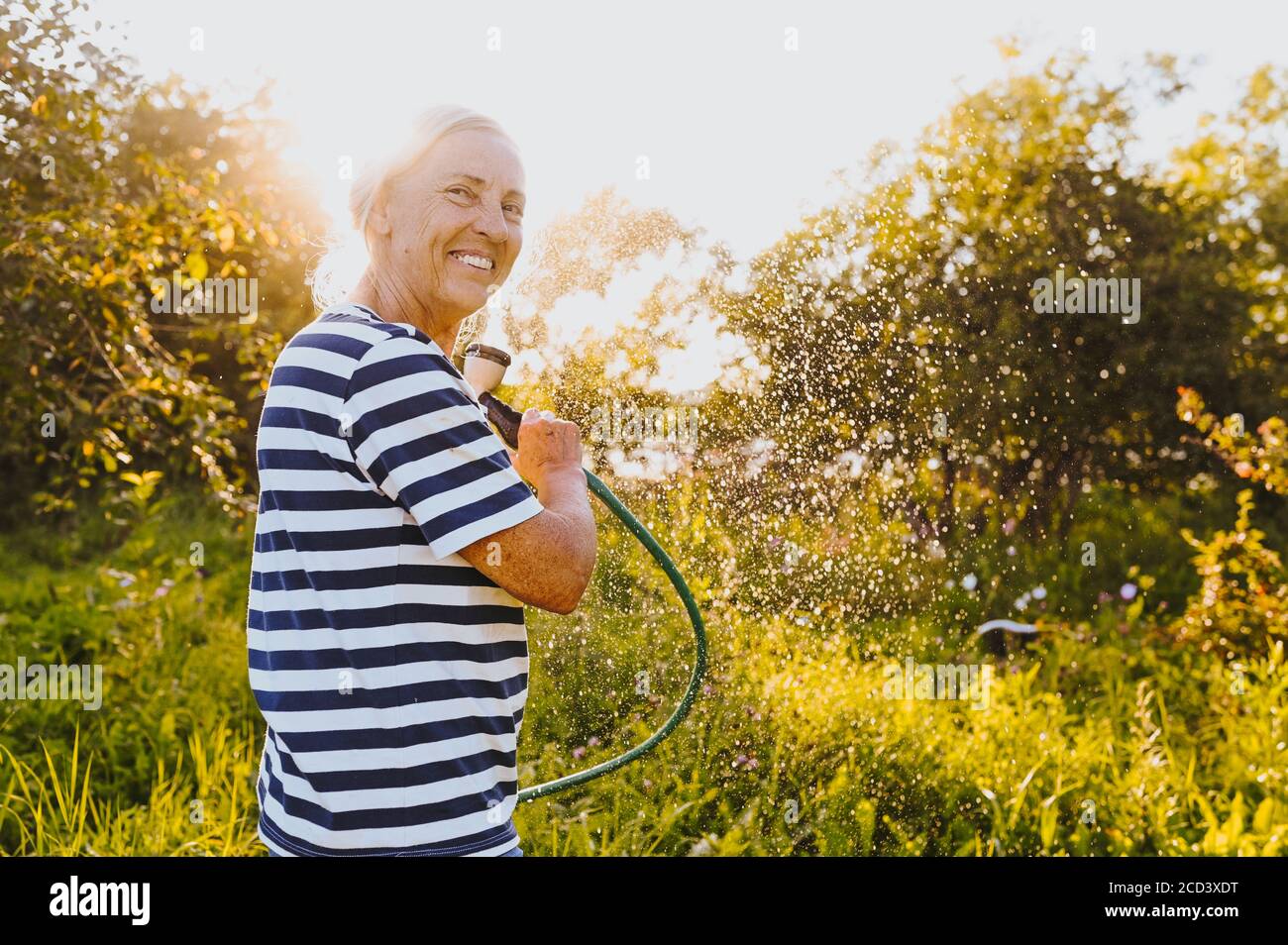 Happy elderly senior woman having fun watering plants with hose in