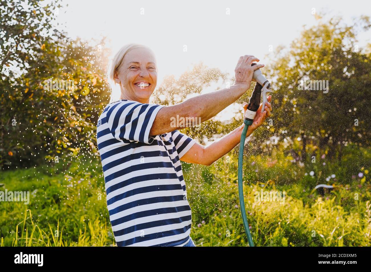 Happy elderly senior woman having fun watering plants with hose in