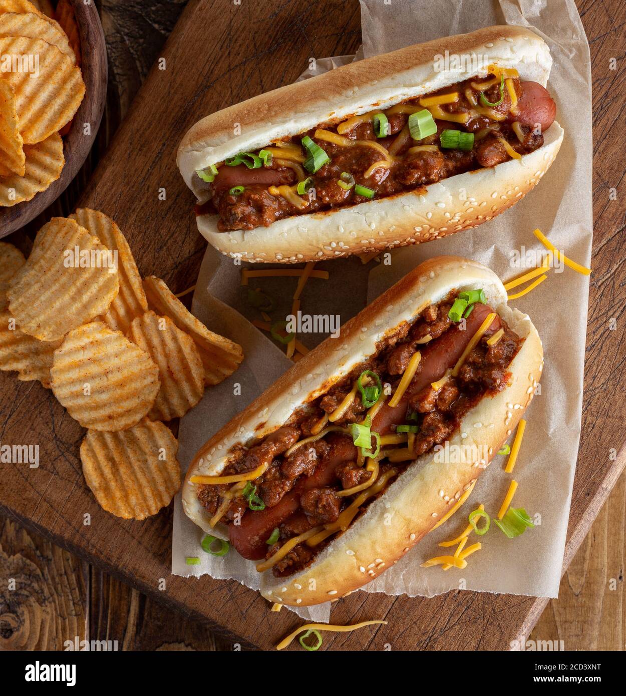 Chili hot dog with cheddar cheese and green onions on a sesame seed bun with potato chips on a rustic wooden board Stock Photo