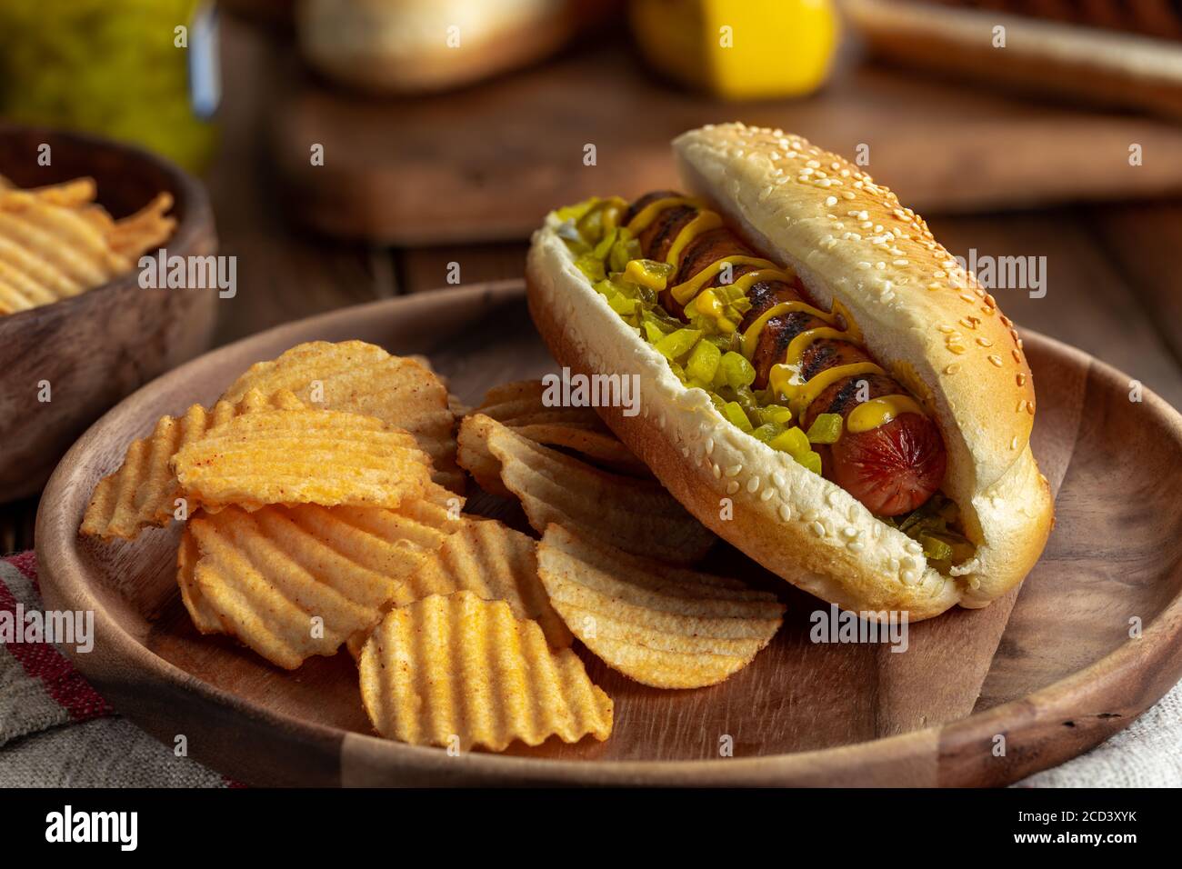 Grilled hot dog with mustard and relish on a sesame seed bun and potato chips on a wooden plate Stock Photo