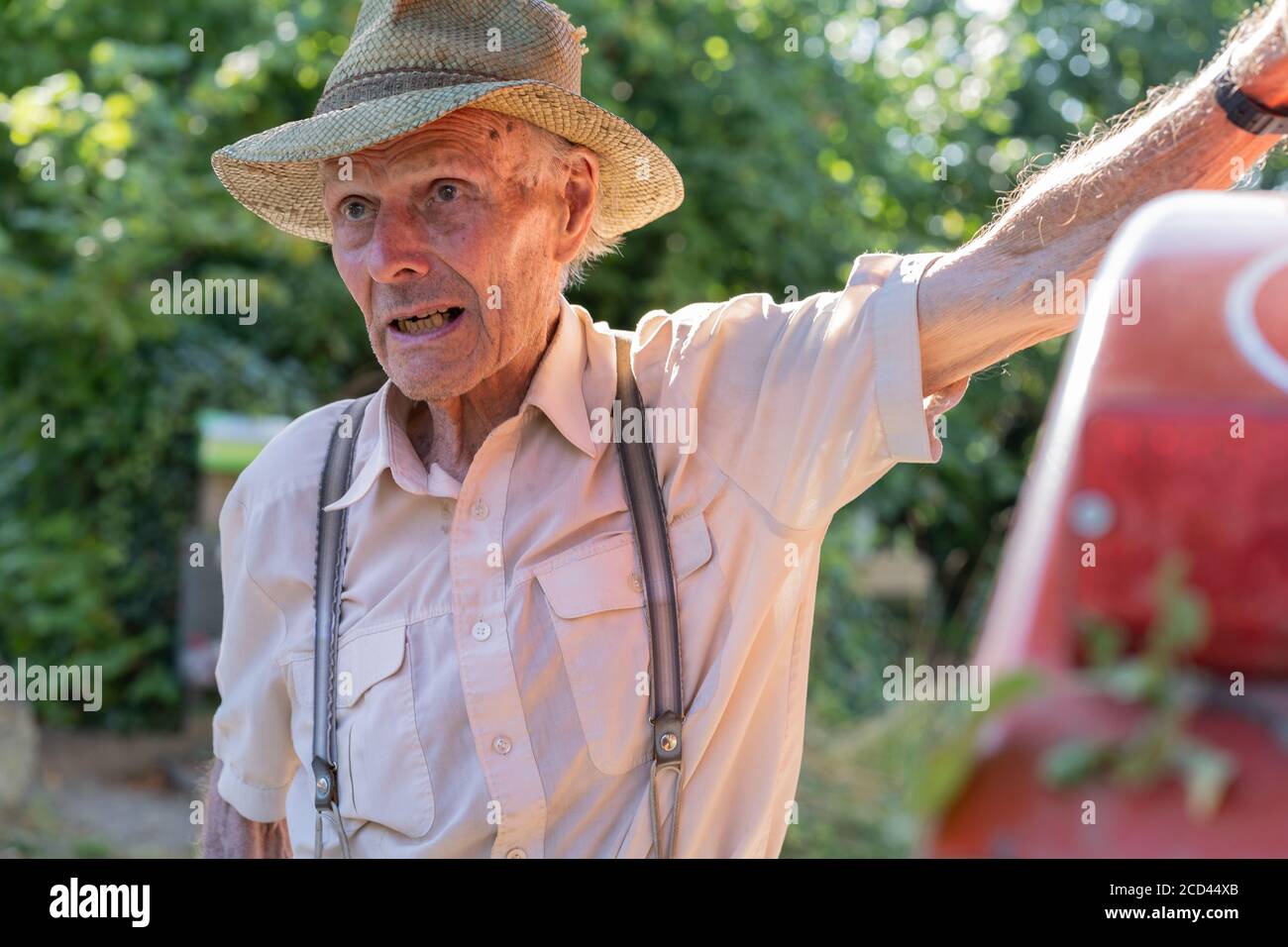 Portrait of very old farmer with straw hat explaining life in front of a red tractor. Real retro, Closeup. Stock Photo