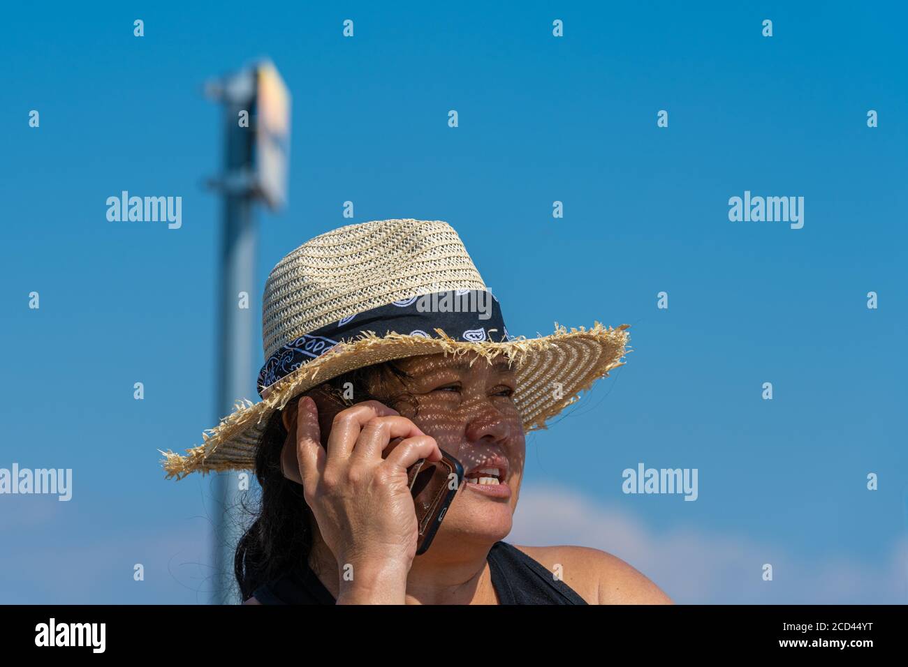 Closeup picture of a middle-aged Asian woman with a straw hat speaking in a smartphone a hot summer day. Bright blue sky in the background Stock Photo